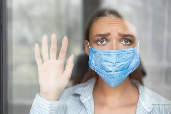 Vrouw dragen beschermende masker kijken uit raam thuis — Stockfoto