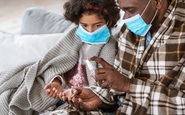 Higiene de manos durante pandemia. Hombre y nieta afroamericanos con máscaras que aplican antisépticos en casa —  Fotos de Stock