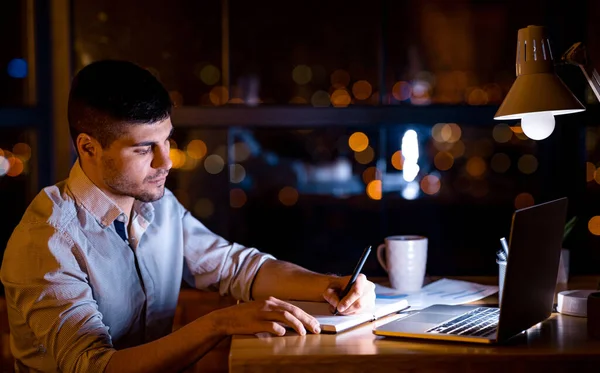 Freelancer Guy prendere appunti seduto al computer portatile che lavora a distanza durante la notte in Home Office — Foto Stock