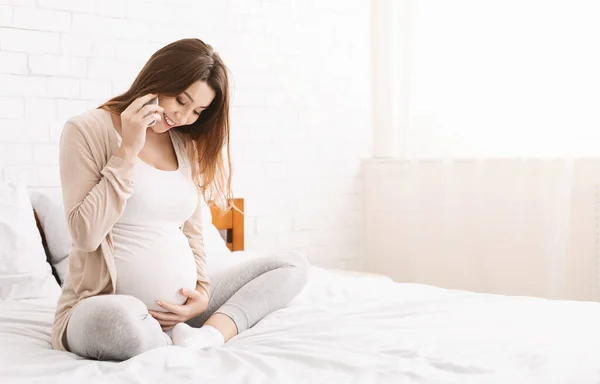 Pregnant woman talking on phone and embracing her belly at home — Stock Photo, Image