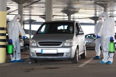 Virüs korumalı giysiler giyen adamlar arabaları kimyasallarla dezenfekte ediyorlar.