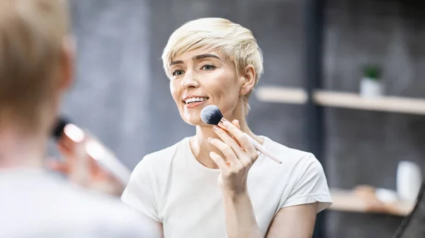 Dame mittleren Alters trägt Gesichtspuder mit Kosmetikbürste in Innenräumen auf, Panorama — Stockfoto