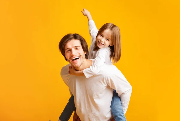 Giocoso bambina seduta sul padre indietro su sfondo giallo — Foto Stock