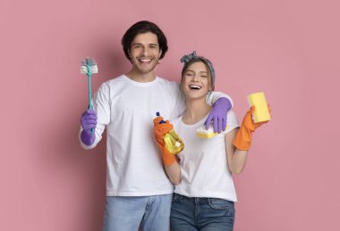 Pembe arka planda elleri temizleme aletleriyle poz veren mutlu çift