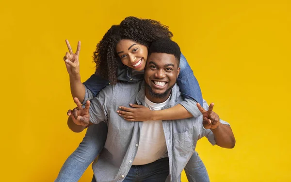 Cheerful black girl piggybacking her boyfriend and showing peace sign — Stock Photo, Image