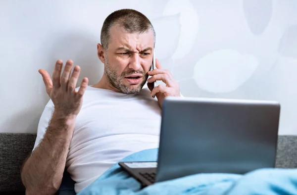 Hombre disgustado hablando por teléfono sentado con el ordenador portátil en la cama —  Fotos de Stock