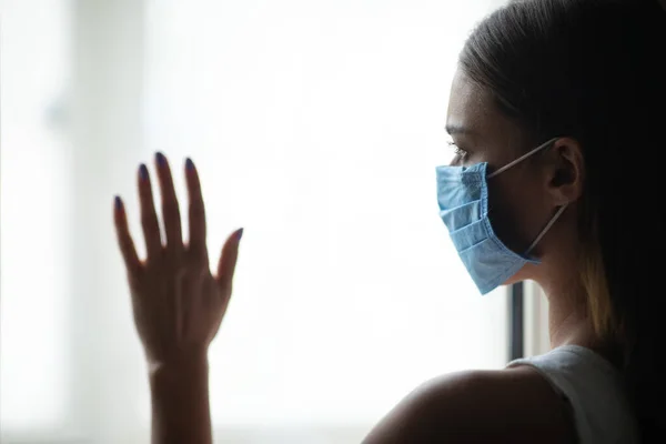 Vrouw in medische masker staande in de buurt van venster verblijf thuis — Stockfoto