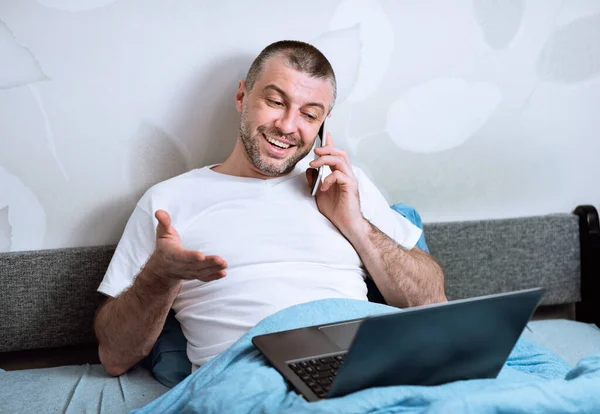 Usmívající se muž mluví na telefonu sedí s notebookem v ložnici — Stock fotografie