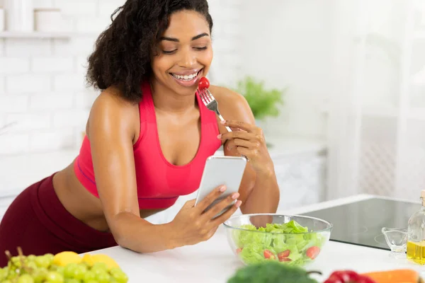 Черная девушка использует смартфон и ест овощной салат — стоковое фото