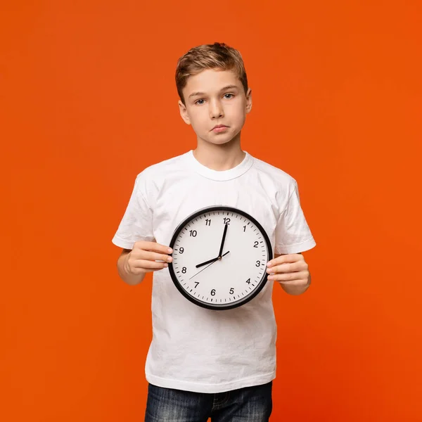Rozrušený dospívající chlapec drží velké hodiny, oranžové studio pozadí — Stock fotografie