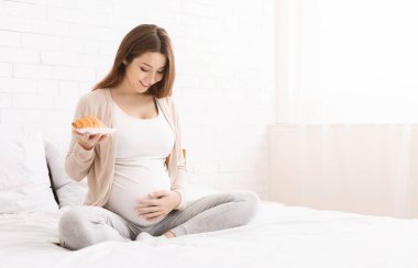 Hamile kadın kruvasan yiyor, yatakta oturuyor.