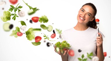 Beslenme ve sağlık. Genç bayan beyaz arka planda kaseden taze salata yiyor. Havadaki sebzelerle kolaj yapıyor.