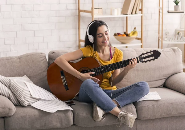 Stay home leisure activities. Happy girl with headphones playing guitar on sofa in living room