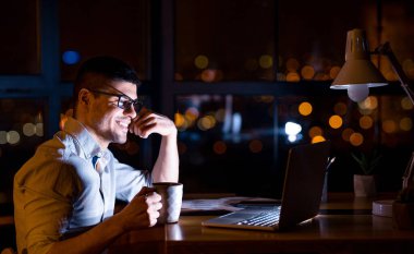 Laptopta oturan adam geceleri ofiste kahve içiyor.