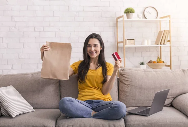 Online winkelen. Jong lachend meisje met laptop, creditcard en papieren tas zittend op de bank thuis — Stockfoto