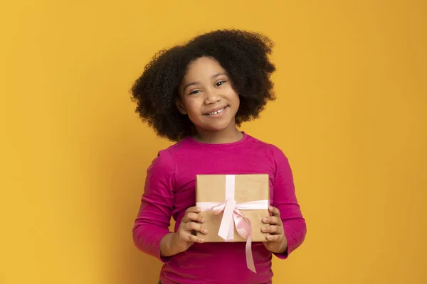 Glimlachend klein zwart meisje holding geschenk doos, poseren over gele achtergrond — Stockfoto