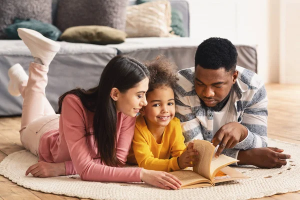 Hija mira a la cámara, padres multirraciales le enseñan a leer —  Fotos de Stock