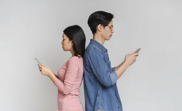 Азиатская девушка и парень стоят спиной к спине, используя мобильные телефоны — стоковое фото