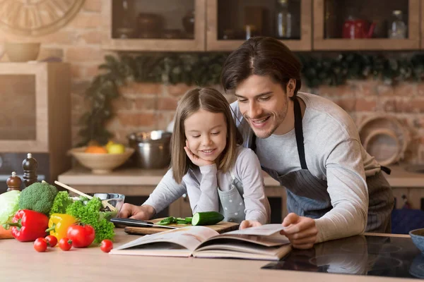Усміхнена маленька дівчинка і її тато перевіряють складові в кулінарній книзі — стокове фото