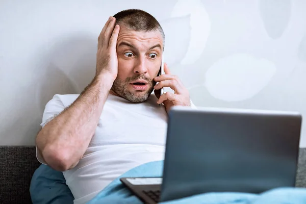 Шокований чоловік розмовляє по телефону за допомогою ноутбука сидячи в ліжку — стокове фото