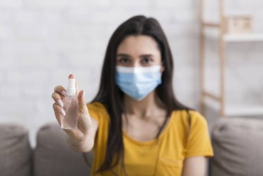 COVID-19 önleme. Koruyucu maskeli kız evde bir şişe dezenfektan tutuyor, seçici odaklı.