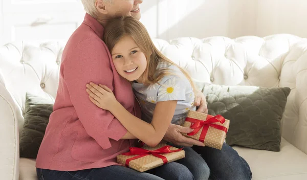 Smiling girl and her grandma with gifts hugging in light room, blank space