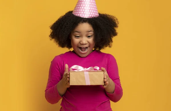 Surpresa de Aniversário. super alegre pouco preto menina no festa chapéu segurando presente — Fotografia de Stock