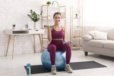 Genç bir kadın evde halterlerle ve spor salonuyla egzersiz yapıyor.