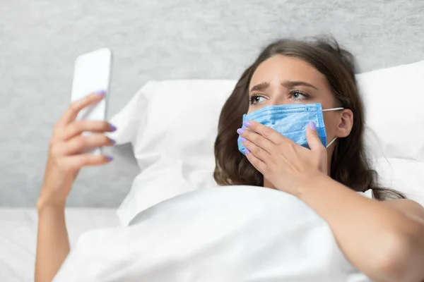 Triest meisje met behulp van smartphone dragen masker liggend in bed binnen — Stockfoto