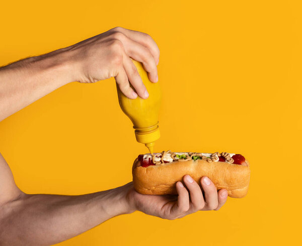 Young man applying mustard on fresh juicy hotdog, orange background. Closeup