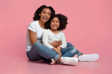 Neşeli bir afro anne ve kızı yerde oturup sarılıyorlar.