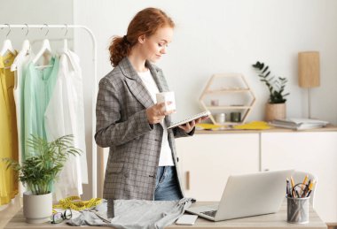 Kadın moda tasarımcısı masasında durmuş kahve içiyor.