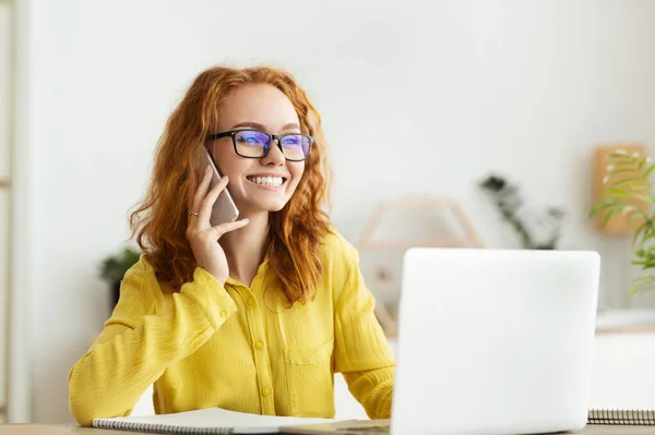 Mujer hablando por teléfono inteligente, trabajando en el ordenador portátil en casa — Foto de Stock