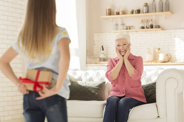 Liten flicka med gåva bakom ryggen och upphetsad mormor i vardagsrummet — Stockfoto