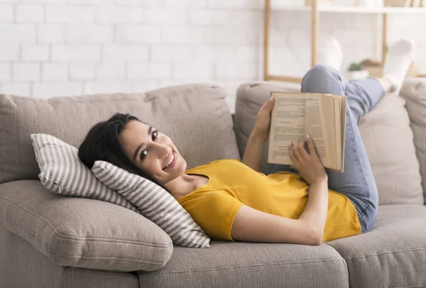Κλειδώστε τις δραστηριότητες αναψυχής. Νεαρή κοπέλα ανάγνωση βιβλίο, ενώ βρίσκεται στον καναπέ στο σπίτι — Φωτογραφία Αρχείου