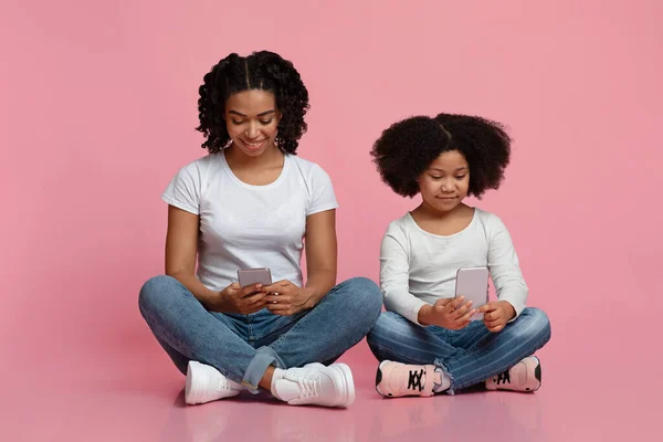 Milenial mujer negra y su hija pequeña sentada en el suelo con teléfonos inteligentes — Foto de Stock