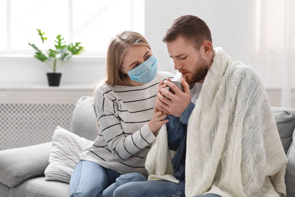 Loving woman in protective mask giving tea to her sick husband