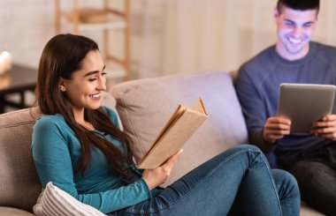 Koltukta oturup kitap okuyan ve tablet kullanan bir çift.