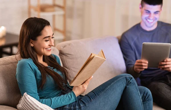 Pár sedí na gauči čtení knihy a pomocí tabletu — Stock fotografie