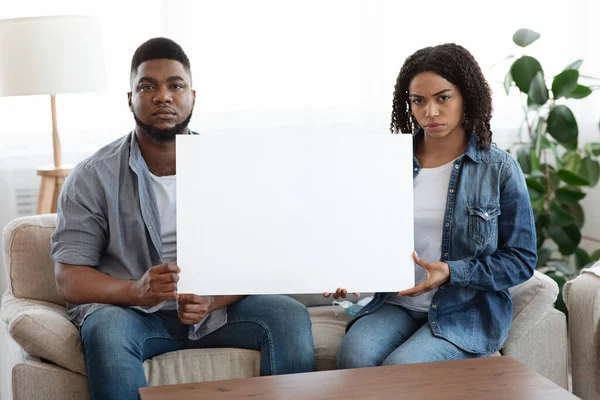 Pareja negra seria sentada en el sofá con pancarta en blanco en las manos — Foto de Stock