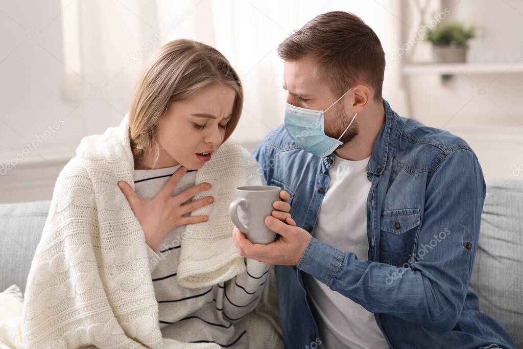 Man in protective mask giving tea to his coughing girlfriend