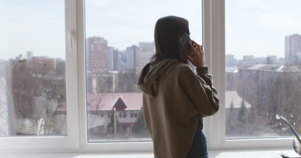 Молодая девушка разговаривает по мобильному телефону, ходит у окна дома — стоковое видео