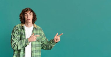 Turkuaz stüdyo arka planında boş bir yeri işaret eden şaşırmış genç adam.