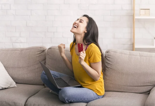 Compras en casa. Emocionado chica latina con tarjeta de crédito y ordenador portátil haciendo la compra en línea en el sofá — Foto de Stock