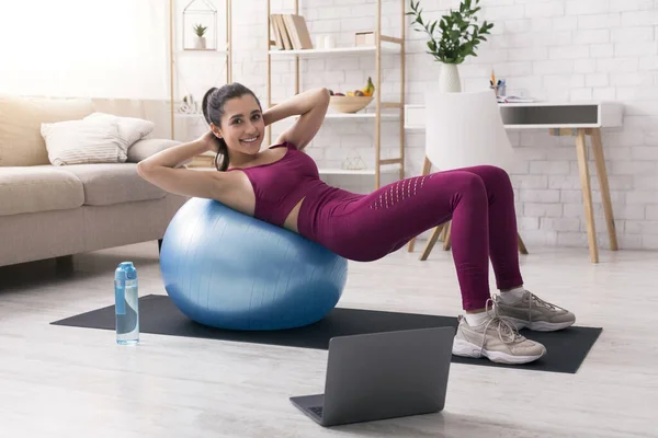 呆在家里，保持活跃。在客厅的笔记本电脑旁，西班牙裔女人带着健身球锻炼 — 图库照片