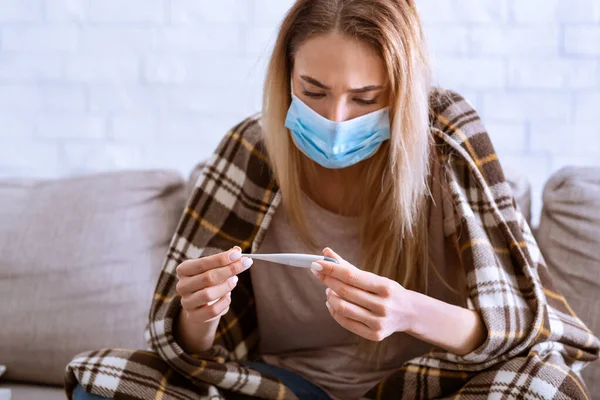 Woman in mask, covered blanket looks at thermometer