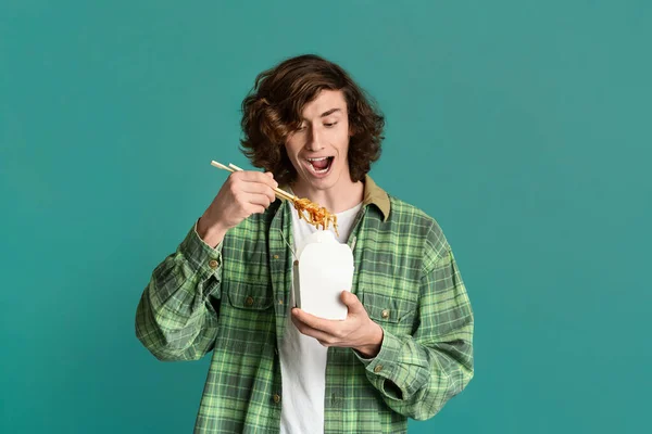 Comida para llevar. Hambriento millennial chico comiendo fideos chinos de la caja desechable en el fondo de color, espacio de copia — Foto de Stock