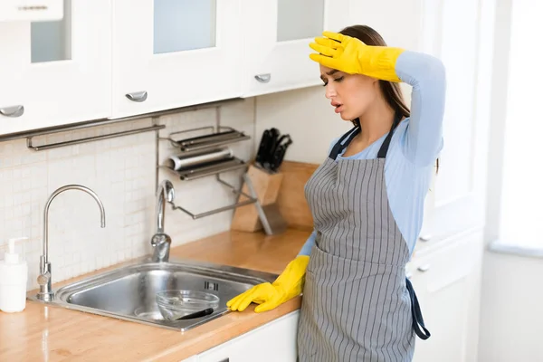 Mujer cansada con guantes de goma mirando platos — Foto de Stock