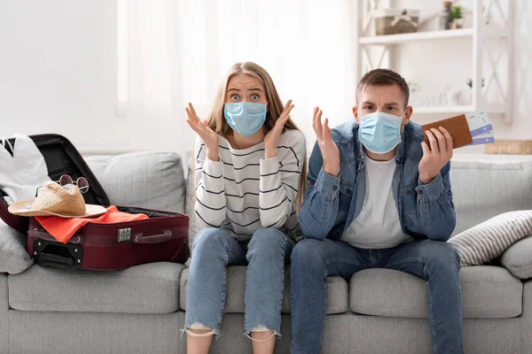 Shocked couple in protective masks sitting with luggage at flight tickets at home — Stock Photo, Image