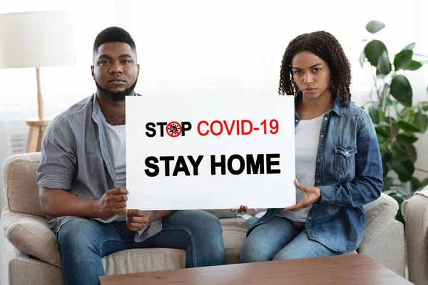 Stay At Home. Black Couple Sitting On Couch With Warning Placard